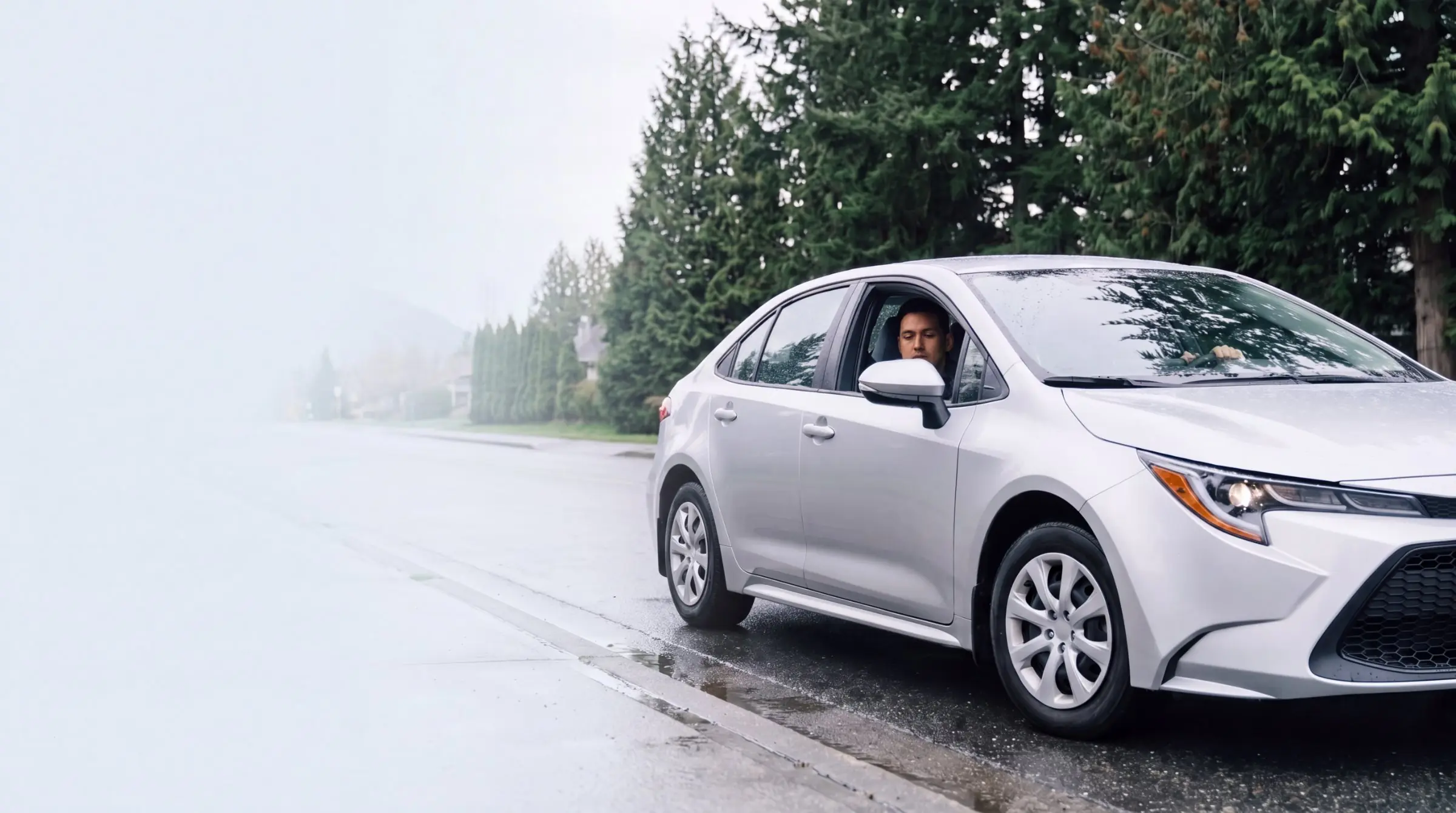 Silver sedan on a rain-wet road, with fog on one side and evergreens behind