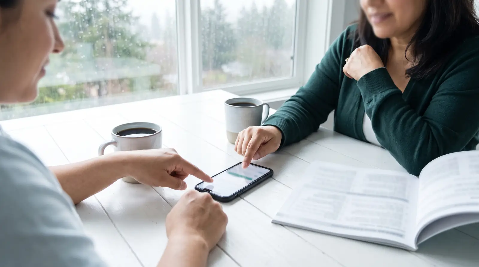 Two women study together at a table, one pointing at a phone beside an open book
